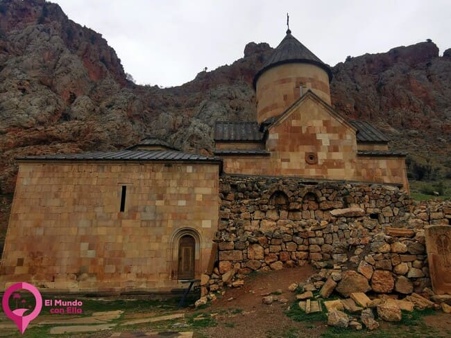 Iglesia de San Juan Bautista de Novarank, destacando su imponente fachada de piedra y su arquitectura medieval armenia, perfectamente integrada con el paisaje montañoso de Vayots Dzor La piedra utilizada en la construcción del Monasterio de Novarank es de un tono cálido dorado, que le da una atmósfera solemne y luminosa a la iglesia, especialmente cuando la luz del sol la ilumina